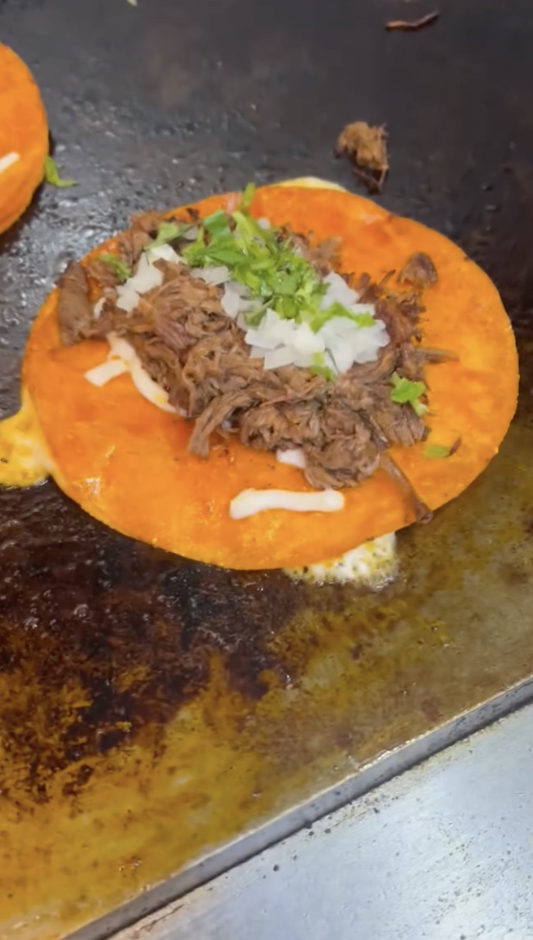 A birria taco on the griddle — red-stained tortilla with shredded beef, onion, cilantro and melted cheese